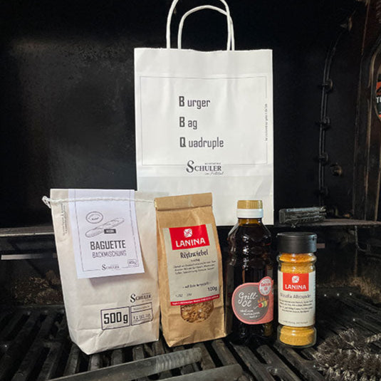 Tüte mit Etikett auf dem BBQ als Burger Bag Quadruple steht, sowie der Tüteninhalt Backmischung Baquette Weizen, Röstzwiebel Packung, Grillöl in Flasche und Gewürzmischung in Gewürzmühle Masshalla Allrounder