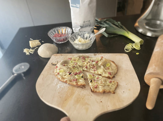 Ein geschnittener Flammkuchen Elsässer Art auf Pizzabrett mit Zutaten im Hintergrund