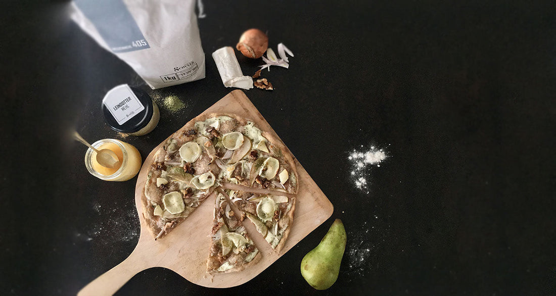 Ein gebackener Flammkuchen auf einem Holzbrett belegt mit Birne, Ziegenkäse, Honig und Walnüssen