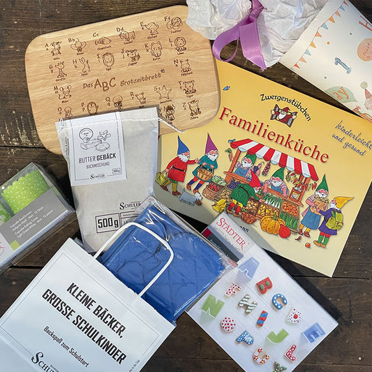 Weiße Tüte mit Logo Kleine Bäcker, große Schulkinder. In dieser Aktionstüte der Kunstmühle Schuler befinden sich eine Backmischung Buttergebäck 500g und 12 Cupcake Backformen, sowie ein Brotzeitbrett abc oder einmaleins, Kinderschürze und -handschuhe in Rosa oder Blau, Zwergenstübchen Back- und Kochbuch und Ausstecherformen Alphabet
