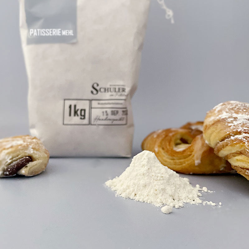 Eine Mehltüte in modernem Design mit einem Haufen Patisseriemehl und Patisserie Stückchen wie Croissant und Plundergebäck