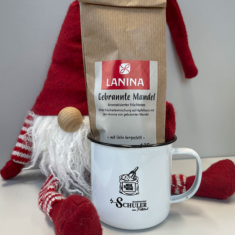 Ein Dekowichtel umarmt einen Emaillen Becher mit Schuler Logo darin befindet sich ein 125 g Tee von Lanina, hier die Geschmacksrichtung Gebrannte Mandel.